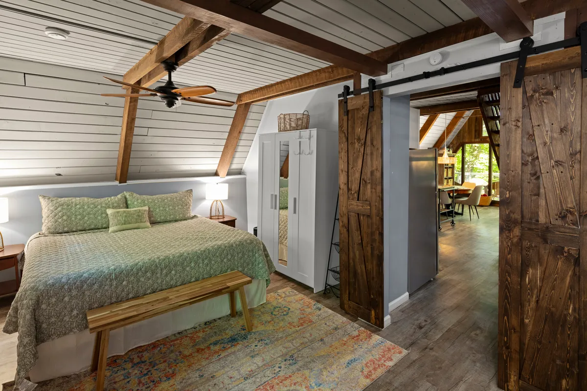 A serene bedroom with green patterned bedding, a wooden bench at the foot of the bed, and a barn-style sliding door. The room is softly lit and the natural tones highlight the timbered ceiling, evoking a peaceful woodland retreat.