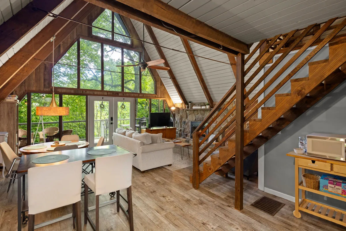 An interior scene with large windows revealing lush greenery, a wooden staircase, a dining table, and a seating area by the fireplace. The setting is ideal for relaxing and enjoying nature views after a day spent skiing or hiking. The open space and practical layout reflect ease of movement from entry to storage areas and main living zones.