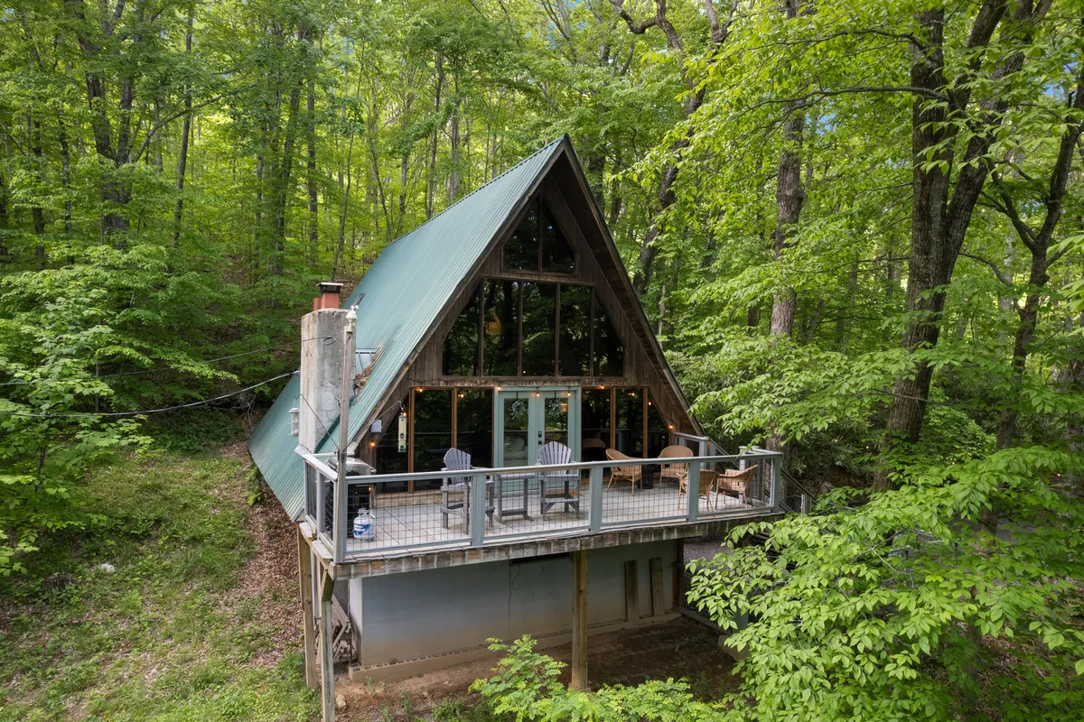 An exterior view of an A-frame cabin with expansive glass windows and a private deck outfitted with comfortable chairs, surrounded by lush forest. The sunlight filters through the trees, highlighting the seamless connection between the indoor living area and the outdoor deck.