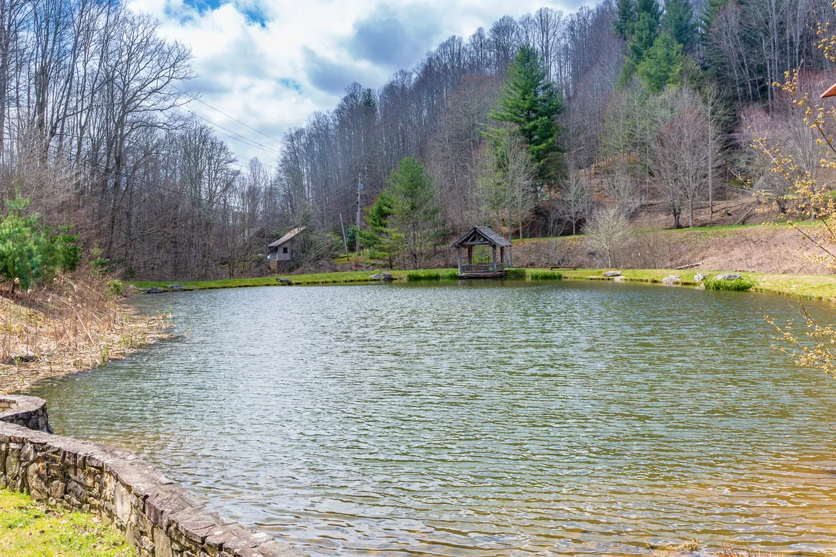 A calm pond surrounded by trees, a wooden gazebo, and a distant cabin. The tranquil setting is perfect for fishing, birdwatching, and quiet moments in nature.