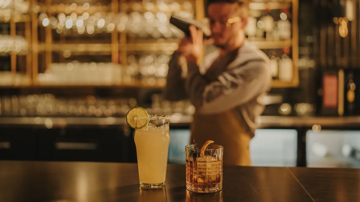 An energetic bar interior with cocktails on the counter and a bartender in action, shaking a drink. The ambiance is lively and sociable, with people enjoying crafted beverages and engaging in conversation.