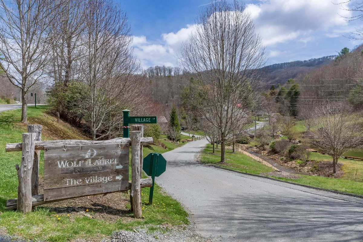 A tranquil exterior scene featuring a wooden signpost at a curve in the road, surrounded by lush trees and landscape. The environment hints at a small village or community, inviting exploration and outdoor activities such as walking, biking, and discovering local artisans.