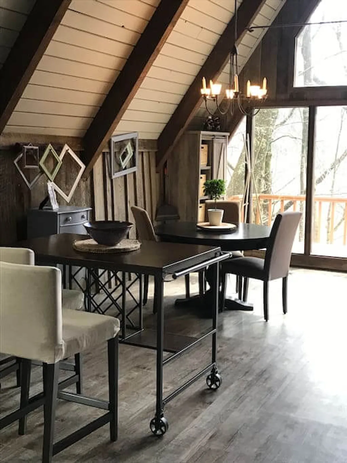 A warm, inviting dining area with a substantial wooden table, decorative chandelier, and expansive windows that look out onto forest views. Wood beams and mirrors add rustic elegance, while the natural light and framed vistas reinforce the cabin’s immersive setting.