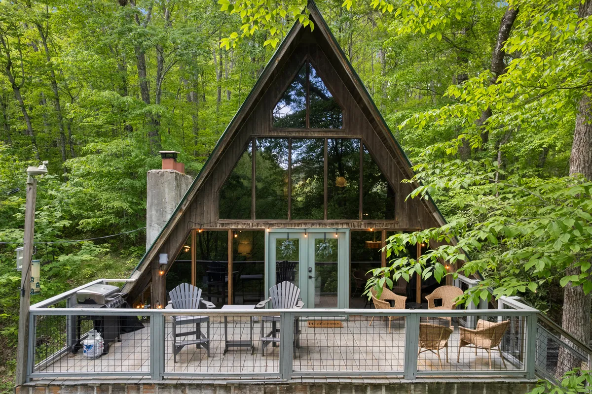 This image showcases a charming A-frame cabin with a striking triangular silhouette, set amid serene North Carolina woods. The cabin features expansive windows that invite sunlight and forest views inside, complemented by a spacious deck furnished for outdoor relaxation. The setting is tranquil, offering unobstructed sunset views from the deck and a cozy, inviting atmosphere that promises peace and connection with nature.