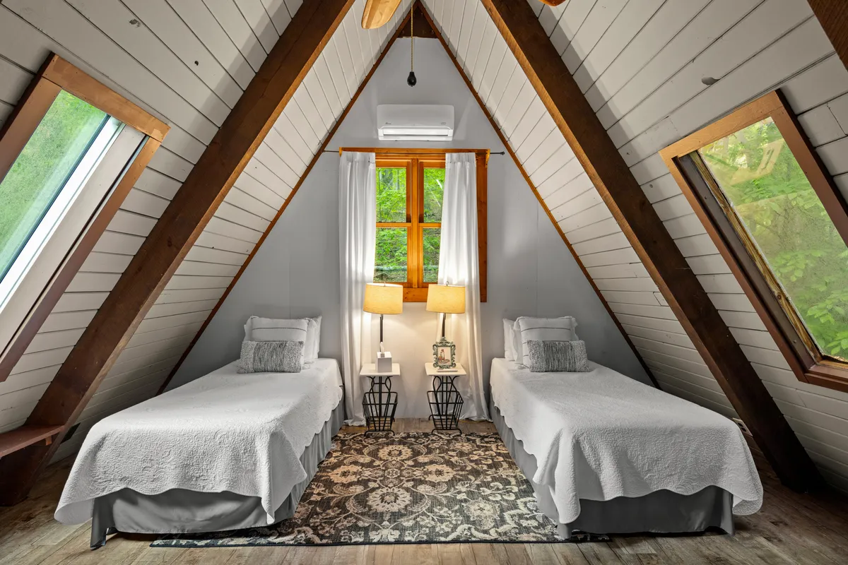 A tranquil bedroom featuring twin beds, a window with curtains that let in natural light, matching table lamps, and a decorative rug. The setting is flexible for families or groups and exudes a calm, restful atmosphere.