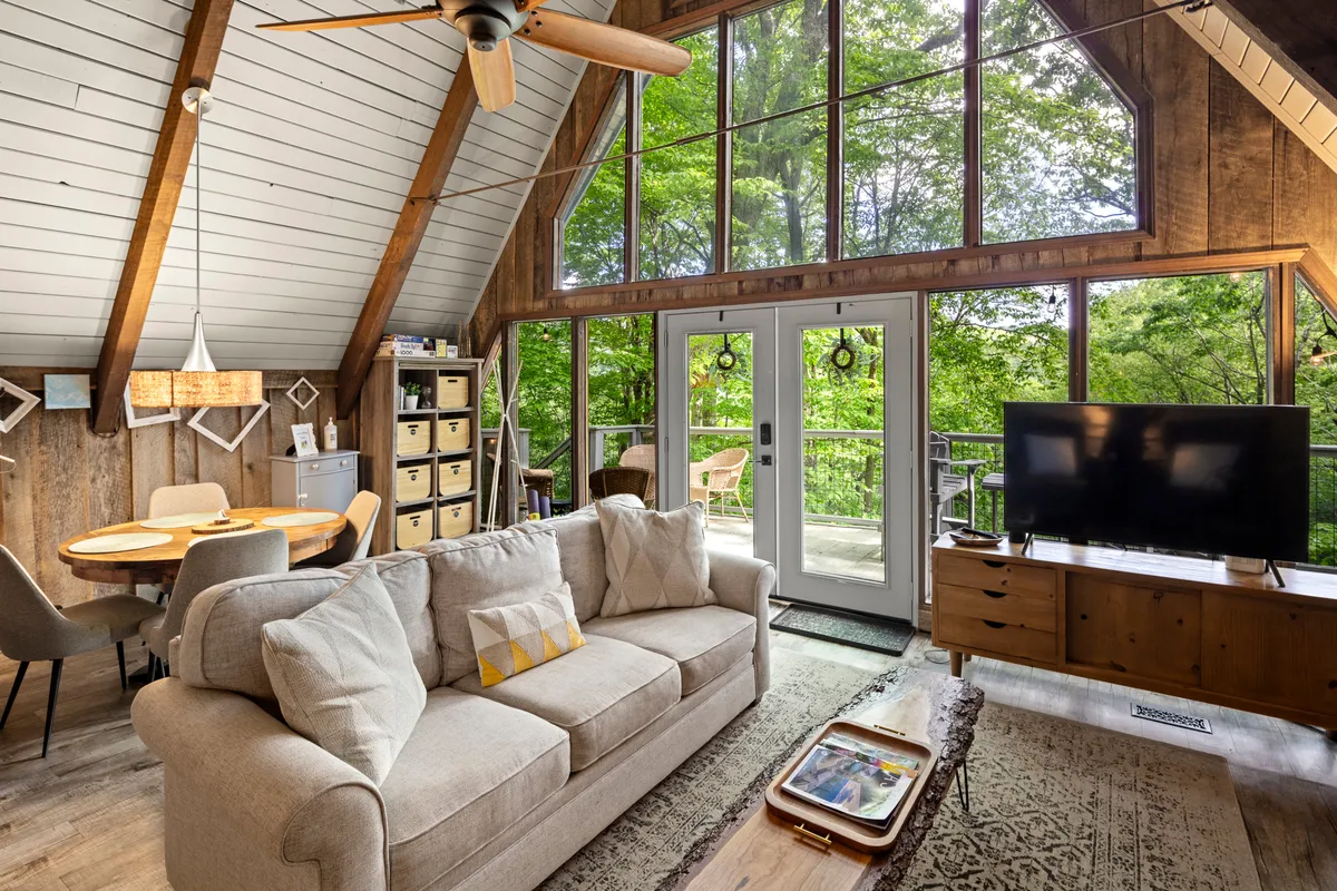 A cozy interior living space featuring a large, comfortable sofa and a dining table, both positioned to take full advantage of sweeping views through floor-to-ceiling windows. Outside, greenery frames the view, reinforcing the blending of interior warmth with natural surroundings.