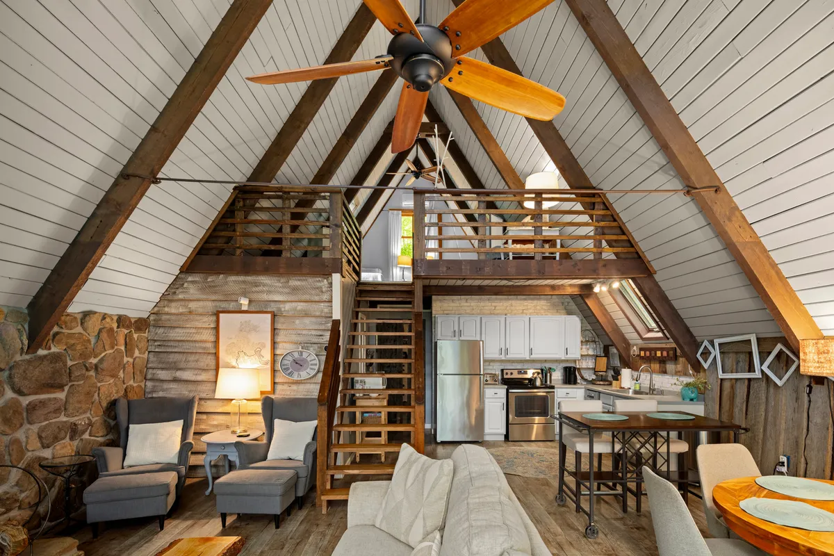 This image showcases a warm and welcoming interior with an A-frame structure, a prominent stone wall, wooden staircase, and a dining area. The scene suggests a cozy living environment ideal for relaxation after a day outdoors. The visible staircase hints at a practical flow from entry to living areas, which supports the transition from adventure to comfort.