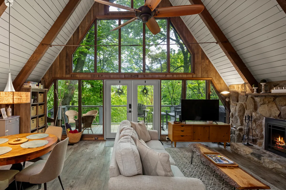 This image showcases a spacious open-concept living area with large windows that allow natural light to flood the space. A stone fireplace creates a cozy focal point, while a comfortable couch and a wooden dining table invite family and friends to gather. Views of the surrounding woodland can be seen through the expansive windows, reinforcing the connection to nature.