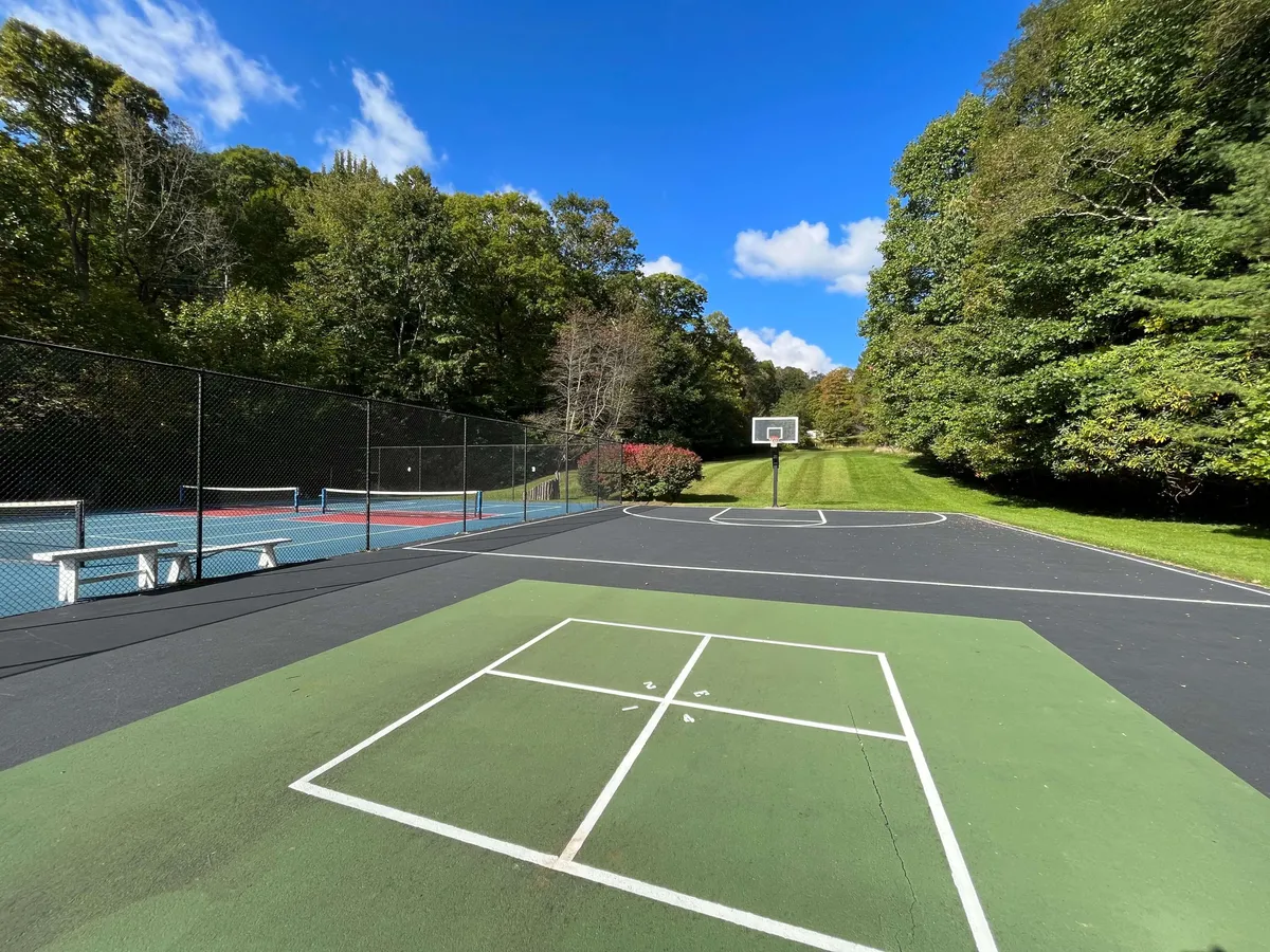 This energetic scene captures the basketball and tennis courts surrounded by lush greenery and blue sky. People are engaged in friendly games, highlighting both tennis and basketball as active, outdoor pastimes. The image conveys a sense of community and togetherness.